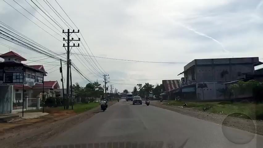 The video shows a street view in Cotpuuk, Indonesia. The scene is a two-lane road with traffic. On the left side of the road, there are houses with red roofs and manicured lawns. There are also utility poles with many wires running across the sky. On the right side of the road, there are more buildings and some vegetation.

Several vehicles are visible, including motorcycles and cars. A cement mixer truck is prominently featured, driving in the same direction as the camera. Other vehicles appear to be traveling in both directions. The weather appears to be overcast with some clouds, and the sky has a visible contrail. The overall impression is of a typical day on a moderately busy road in a semi-urban or rural area. There is no discernible emotional tone.

No text is clearly legible within the video frames. The time of day appears to be daytime, likely late morning or early afternoon, given the lighting conditions. The presence of houses, shops, and a main road suggests a populated area.