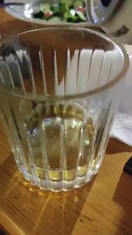 The video shows a person pouring a light-colored liquid from a silver container into a clear, textured glass. The glass is about a third full. The liquid appears to be creating some foam as it enters the glass. The glass sits on a wooden table, next to a white napkin and a small black device. A plate of salad is partially visible in the background.