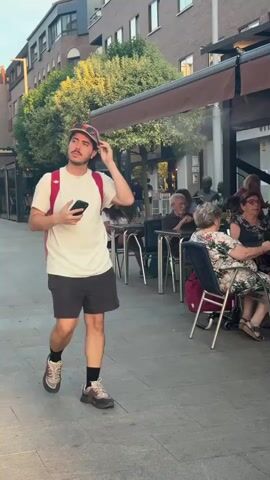 A man walks on a sidewalk past an outdoor cafe. He is wearing a white t-shirt, black shorts, a baseball cap with sunglasses perched on top, and a red backpack. He holds a smartphone in his right hand and adjusts his cap with his left. Several people are seated at tables in the background. The sun is setting, casting long shadows.
