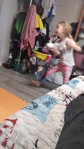 A young girl with blonde hair in pigtails, wearing a white t-shirt with a graphic and colorful patterned leggings, is standing in a cluttered room and holding a stuffed animal. She is barefoot and appears to be in motion. The room contains a variety of items including clothes hanging on a rack, several glass jars, a yellow plastic basin, and stuffed animals on the floor. A bed with a patterned comforter is visible in the foreground. The floor is a mix of grey laminate and an orange puzzle mat.