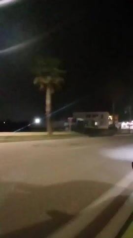 The video depicts a street at night, with a building visible on the left and streetlights illuminating the area. Cars pass by on the road, their headlights creating bright streaks. The overall scene is somewhat blurred due to the low light conditions and possible movement during the recording.