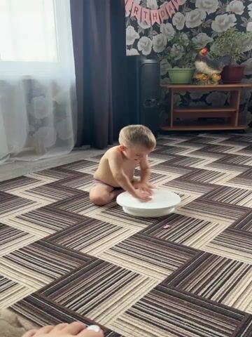 A young child, likely a boy, is kneeling on a patterned rug in what appears to be a room. He is shirtless and wearing striped shorts. In front of him is a white, circular cake stand, and he is placing his hands on it, seemingly engaged in play or exploration. The background shows a window with sheer curtains, a dark brown curtain, a black speaker, and a shelf with a potted plant and a decorative chicken. The wall behind the shelf is covered in floral wallpaper. A banner with the words "HAPPY BIRTHDAY" is visible at the top of the background. The lighting suggests it is daytime, with sunlight coming from the window. The child appears focused on his interaction with the cake stand.