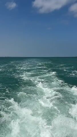 The video shows a wake from a boat moving through the ocean. The water is a deep green, and the wake is a frothy white with churning waves. The sky is a clear blue with a few scattered white clouds. In the distance, land can be seen on the horizon. The camera is positioned behind the boat, looking out over the water. The video captures the motion and energy of the boat's passage through the sea.