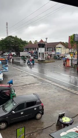 The video shows a street scene with a roundabout and several vehicles. It is a cloudy day, and the road appears wet, suggesting it may be raining or has recently rained. In the foreground, a black car is parked on the left side of the road. A blue bus is also visible on the left, and several tuk-tuks are present throughout the scene. A traffic light stands in the center of the roundabout. People are seen walking with umbrellas, and some are riding motorcycles. Buildings line the street on the right side, with various signs and shops visible. A tall antenna tower can be seen in the background on the left. The overall impression is of a busy street in a tropical or subtropical region experiencing inclement weather.