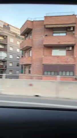 This video shows a view from inside a vehicle traveling along a road. The camera captures a series of apartment buildings lining the right side of the road. The buildings vary in height and color, with some featuring balconies and awnings. Trees and greenery are visible between the road and the buildings. The road appears to have at least two lanes, separated by a concrete barrier. Other vehicles, including a white car, are also seen on the road. The sky is visible above the buildings, suggesting it is either morning or late afternoon. The motion of the camera indicates the vehicle is in motion, possibly on a highway or a major road.