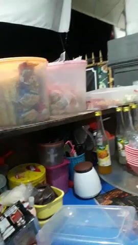 The video shows a cluttered shelf with various containers and bottles, some of which appear to contain food items. In the foreground, a blue plastic container is visible. The lighting suggests an indoor or covered outdoor setting, possibly a food stall or a kitchen. The scene is somewhat out of focus and dimly lit.