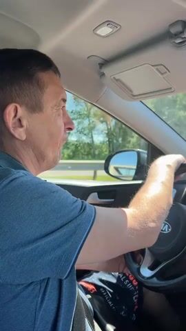 A man is driving a car on a sunny day. He is wearing a blue t-shirt and has short brown hair. The camera is positioned inside the car, looking at the man from the side. The man turns his head and smiles at the camera. He then turns his attention back to the road. The car appears to be on a highway, with trees and a blue sky visible outside the windows.