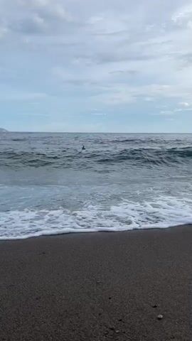 The video shows a view of a beach with dark sand and the ocean. Waves are breaking on the shore, creating white foam. In the distance, there are several people in the water, some appearing to be swimming or possibly surfing. The sky is cloudy. The scene is a tranquil coastal environment.