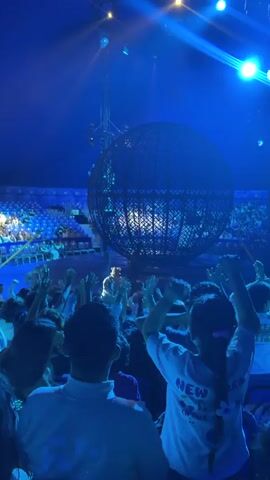 This is a video taken from the perspective of an audience member at a circus performance. The audience is looking towards a large, spherical cage. Inside the cage, a performer is visible, and they are manipulating a glowing, multicolored ball. The audience members, including the person recording, have their arms raised in the air, suggesting excitement or participation. The environment is dimly lit, with bright spotlights illuminating the performance area. The overall atmosphere appears to be lively and engaging.