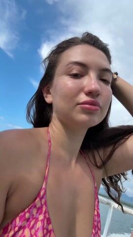 The video shows a woman on a boat, wearing a pink floral bikini top. The water is blue and the sky is partly cloudy. She looks into the camera briefly before looking down. The video cuts to a woman in sunglasses and then cuts again to another woman on the boat in sunglasses, wearing a dark-colored top. There are other people on the boat.