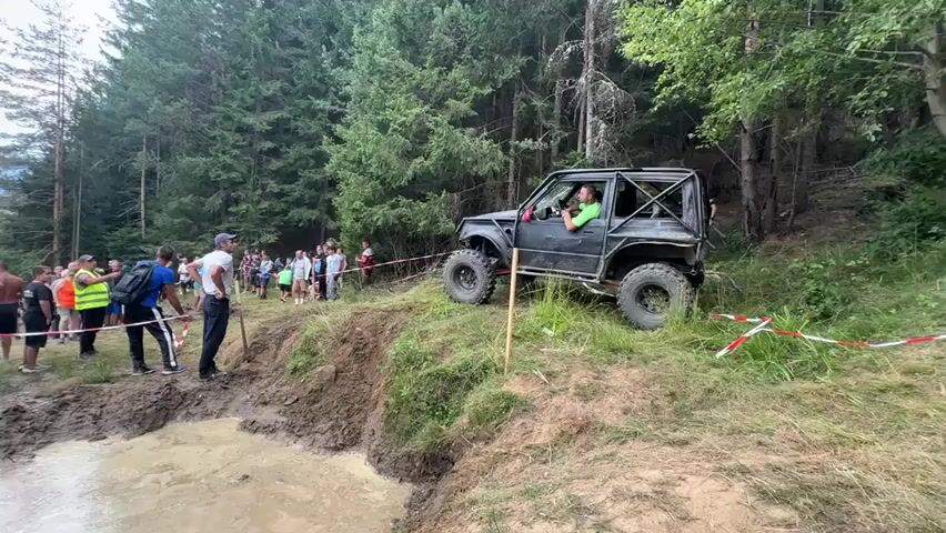 A group of people are watching an off-road vehicle navigate a muddy obstacle course in a forest. The vehicle, a dark-colored Suzuki Samurai with a roll cage, is positioned on a grassy incline next to a muddy pit. A person in the driver's seat appears to be signaling. Several spectators stand behind a red and white caution tape barrier on the left side of the frame, facing the vehicle. The environment is a dense forest with tall trees. The lighting suggests it is daytime.
