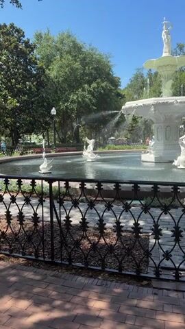 This video shows a beautiful, multi-tiered white fountain in a park on a sunny day. The fountain features several white sculptures, including a figure at the very top and several smaller figures surrounding the lower tiers, with water spraying from various points, creating a refreshing and lively atmosphere. The fountain is set in a pool of water, and the surrounding area is lush with green trees, indicating a pleasant outdoor setting.

In the foreground, a decorative black metal fence with an intricate scroll pattern separates the viewer from the fountain and pool. Beyond the fence, a paved walkway made of reddish bricks is visible, with shadows suggesting a bright, sunny day. The background reveals a dense canopy of green trees, typical of a park or garden. The sky is a clear, vibrant blue, further reinforcing the impression of good weather.

Although no people are prominently featured in the immediate vicinity of the fountain, hints of activity can be seen in the distance, suggesting that this is a public space where people are present. The overall mood conveyed by the scene is peaceful, serene, and idyllic, enhanced by the sight and sound of the flowing water and the natural beauty of the park. The time of day appears to be midday, given the bright sunlight and the sharp shadows. The presence of mature trees and the style of the fountain might suggest a historical park setting. The location is identified as Savannah, United States of America. No text is visible in the media file.