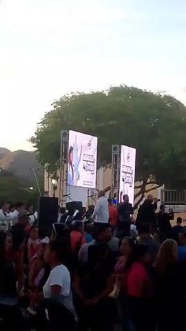 The video shows a daytime outdoor event with a crowd of people gathered in front of a stage with two large video screens. The screens display a poster with a woman's image and the words "MEMORIA CULTURAL." Some people are standing on the stage, appearing to speak or perform. A large tree is behind the stage, partially obscuring a building. Mountains are visible in the distance. The crowd seems to be watching the stage, and some individuals are possibly interacting with each other.