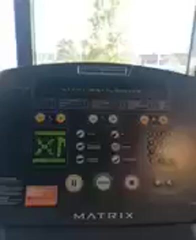 The video shows a close-up of the control panel of a Matrix treadmill. The screen displays text in French, which appears to be instructions or a prompt. The screen also shows a graphical representation of a heart rate monitor. Various buttons are visible, including a pause button, a start button, and buttons for selecting workout programs. An orange button labeled "go" is also present. The treadmill is set up indoors, with a blurred view of a window showing trees and cars outside. The overall lighting suggests it is daytime. The camera is relatively steady, with slight movements.