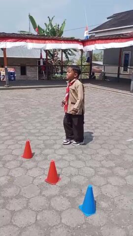 A young boy in a scout uniform walks around two orange traffic cones in an outdoor paved area. The boy is wearing a tan uniform shirt with a red and white scarf, dark pants, and white sneakers. He is looking to his left and appears to be moving in a clockwise direction around the cones. In the background, there is a covered structure decorated with red and white fabric, and trees. The sky is clear and bright. The video cuts off as the boy is passing the second cone.