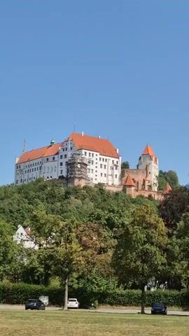 The video shows a large, white castle with a red-tiled roof situated on a hill covered in lush green trees. The castle appears to be undergoing some renovations, with scaffolding visible on one section of the building. In the foreground, there is a grassy field with several cars parked along a road that runs parallel to a tree line. The sky is clear and blue, indicating a sunny day. The overall scene depicts a picturesque view of a historic landmark surrounded by nature.