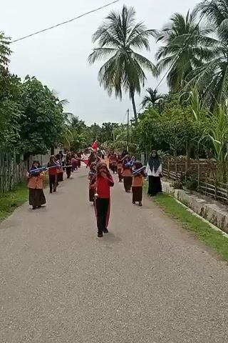 A marching band composed of young girls in brown uniforms and hijabs walks down a paved road lined with palm trees and houses. The girls carry blue and white props, possibly mock instruments or batons, and some hold small flags. A girl in a red and black outfit, also wearing a hijab, leads the band, holding a baton and a flag. Another woman in a black hijab stands on the side of the road watching. The sky is overcast.