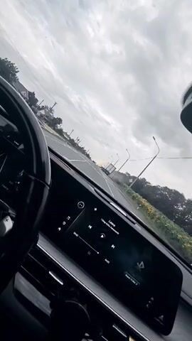 A first-person perspective video captures the interior of a car. The dashboard features a large touchscreen display showing music controls and song information that reads "Lalala" and "The Black Eyed Peas." To the left, a portion of the steering wheel is visible. The view through the windshield shows a road with white lane markings, a cloudy sky, and trees lining the right side of the road. In the distance, a white truck is driving on the road. The camera appears to be mounted inside the car, possibly on the dashboard or windshield, and is angled slightly upwards. The video has a duration of 4 seconds.