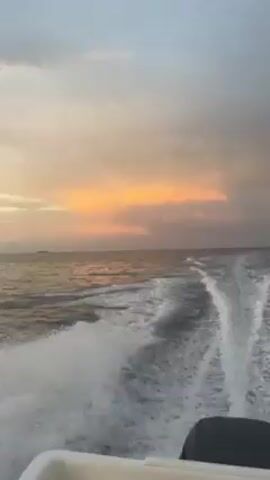 The video shows a boat moving through the water, creating a visible wake. The sky is a mix of grey and orange hues, suggesting either sunrise or sunset. In the distance, a landmass is visible on the horizon. The camera angle changes, revealing parts of the boat, including ropes and a fishing pole. The water appears choppy.