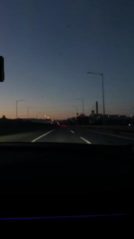 The video shows a first-person view from inside a car driving on a highway at dusk or dawn. The sky is a deep blue fading to orange near the horizon. Streetlights and distant lights from buildings are visible along the sides of the road. The car's dashboard is in the foreground, with a dark silhouette of the car's interior. White lines mark the lanes of the highway. The road appears to be relatively clear, with a few other vehicles visible in the distance.