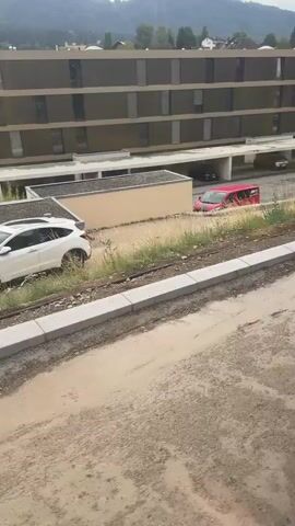 A red van is shown driving through a flooded area in front of a building. The water level is high enough to be covering the van's wheels and lower body. The van appears to be slowly moving forward. The environment is muddy and appears to be undergoing construction, with piles of dirt and construction materials visible. The building behind the flooded area is a multi-story structure with a dark facade and numerous windows. Mountains are visible in the background. The scene suggests a recent heavy rainfall or a drainage issue.