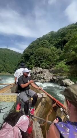 A group of people are on a boat in a river, surrounded by lush green mountains under a blue sky with white clouds. A man in a black t-shirt and shorts is using a long pole to steer the boat through the rushing water. He is wearing a white hat and sunglasses. Another person wearing a white t-shirt is visible in the background, also on the boat. In the foreground, parts of other passengers are visible, including a person with a pink bow in their hair and another person holding a phone to take a picture. There is a sign on the side of the boat with Japanese writing. The boat appears to be moving forward through a rocky section of the river.
