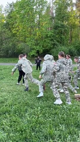The video shows a group of around 10 young people in camouflage clothing running across a grassy field. They are participating in what appears to be a military-style exercise or training activity. An individual sits in a chair, and the group initially runs past them before turning towards some obstacle in the distance. Trees surround the field, and the sky is blue.