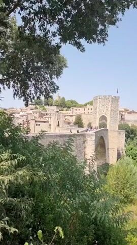 The video pans across a scenic landscape featuring a river and bridges. In the foreground, lush green foliage partially obscures the view. The first bridge, made of stone, has an arched structure and a prominent tower with flags at its peak. Several people are visible walking on this bridge. Beyond the stone bridge, the camera moves to reveal a wider view of the river, surrounded by dense greenery and trees. In the mid-ground, a modern concrete arch bridge spans the river, its structure a stark contrast to the ancient stone bridge. In the background, rolling hills covered in trees and vegetation create a soft horizon, under a clear blue sky with a few wispy clouds. The overall impression is one of historical and natural beauty.
