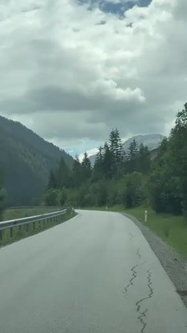 This media file captures a scenic drive through a mountainous region, likely Flachauwinkl, Austria. The scene is dominated by a winding asphalt road cutting through a lush green valley. Towering, verdant mountains rise steeply on the left, densely covered in trees. On the right, the road is bordered by a metal guardrail and then a slope covered in trees and grass, with a few scattered buildings or structures visible higher up the mountainside. A line of tall, dark green pine trees flanks the right side of the road, leading the eye towards the misty, cloud-covered peaks in the distance.

The sky is overcast with dramatic, grey clouds, suggesting either an approaching storm or a generally cloudy day, likely in the afternoon given the diffused light. The overall mood is one of peaceful solitude and natural beauty, with the empty road inviting exploration.

There are no people visible in the media. The primary objects are the road, the guardrail, the trees, and the mountains. The setting is clearly a mountainous, forested area, indicative of a rural or alpine location. The activity taking place is a drive along this scenic route. Notable details include the cracked asphalt on the road's surface, suggesting wear and tear, and the white roadside markers. No visible text is present.