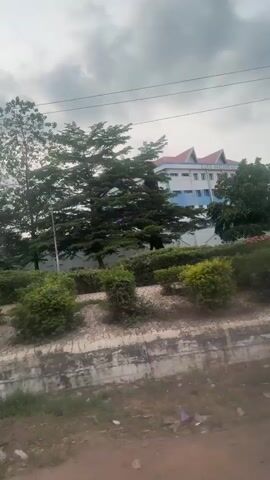 The video shows a street view from a moving vehicle. There are trees and bushes along the side of the road, with buildings visible in the background. The sky is cloudy. As the vehicle moves, the buildings and surrounding landscape change. Eventually, the vehicle approaches a gate with distinctive architectural features.
