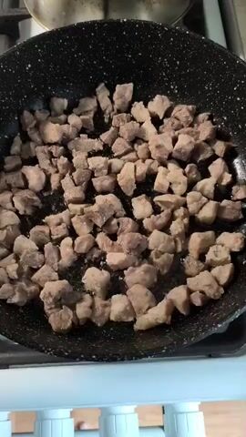 In the video, diced pieces of meat are being fried in a black non-stick pan. The meat is cut into small cubes, roughly 1-2 cm in size, and appears to be lightly seasoned with salt and pepper. The pan is on a stovetop, indicated by the white knobs and the edge of a white appliance visible at the bottom of the frame. There is a small amount of cooking oil in the pan, and the meat is sizzling as it cooks, indicating it is in the process of browning. Some pieces of meat have begun to brown on the edges. A pot with a shiny surface is partially visible in the upper left corner of the frame. The lighting appears to be from overhead. The video is a close-up shot, focusing primarily on the food being cooked.