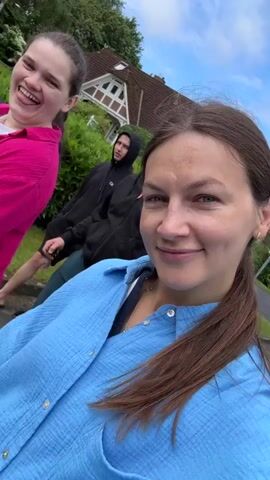 A young woman with long brown hair, wearing a light blue button-down shirt over a dark top, films a selfie video, smiling at the camera. Behind her to the left, another young woman with a long braided ponytail, wearing a pink top, laughs. Further back, three individuals are visible: a young man in a black hoodie and shorts, a man in a black long-sleeved shirt, and a young woman in a dark vest and white top. They appear to be outdoors on a street with a house and green foliage in the background under a cloudy sky. The woman filming makes a funny face at the camera at the end of the video.