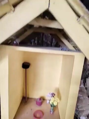 The video shows a small, open-fronted structure with yellow walls. Inside, on the floor, there is a pink dish with some residue and a round pink container. To the right, a vase holds a bouquet of flowers in shades of yellow, white, and purple. To the left, a thin red pole with a black cap stands upright. The environment appears somewhat dusty and neglected, with some debris visible on the ground outside the structure. The camera movement is shaky, suggesting it may be handheld.