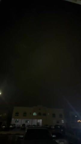 The video shows an exterior view at night. A tan-colored building is partially visible at the bottom of the frame, with several arched windows and a central arched entrance.  A line of parked cars is in front of the building. The sky is dark, nearly black, and it obscures most of the view. Brief flashes of light streak across the sky. The camera angle is low, looking upward. A dark, flat surface, possibly a part of a building, is visible at the top of the frame, partially obscuring the sky.