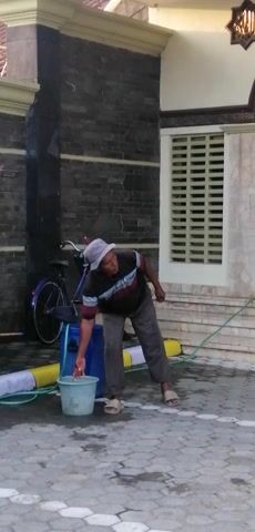 A man wearing a hat and a striped shirt is seen in a courtyard, leaning over a bicycle. He is near two buckets and a yellow object. The ground is paved with gray tiles, and a wall with a large window is visible behind him.