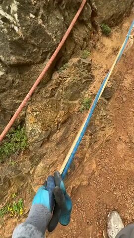 The video opens with a first-person perspective shot of a pair of gloved hands holding a blue and yellow rope. The camera tilts upward to reveal a person in a red jacket climbing up a steep, rocky hillside using two ropes anchored along the path. The ground is mostly reddish-brown dirt and scattered rocks, with some green vegetation interspersed. The camera follows the climber briefly, then pans to a wider shot of the hillside, showing the ropes extending upwards towards a hazy sky. Finally, the camera turns to face a woman in a gray sweatshirt and black fanny pack. She is standing on a green grassy area, with rocky cliffs and the ocean visible in the background. She looks directly at the camera.