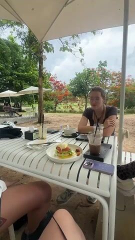 The video shows two people seated at a white table with a white umbrella overhead, enjoying a meal outdoors. A woman with glasses is sitting at the table, looking off to the side and talking. In front of her is an iced coffee, a phone and a plate with food. On the plate is a stacked dish, likely an egg benedict with greens, salmon and tomatoes. The person filming is seated at the table, with their legs visible in the foreground. The camera zooms in on the plate of food. In the background are other tables with people, trees and a cloudy sky. The environment appears to be a cafe or restaurant with outdoor seating.