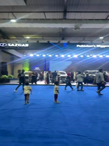 The video shows an indoor exhibition hall with a large blue carpet. There is a stage or display area in the background with several new cars, some with lights. A banner above the cars reads "Pakistan's Biggest NEV Lineup." People are milling around the cars, looking at them. In the foreground, two young children are standing on the blue carpet, facing away from the camera. The environment appears to be well-lit, with overhead lighting and spotlights. The overall atmosphere suggests a car show or product launch event.