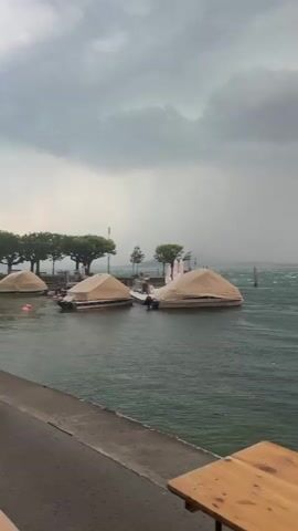 The video captures a stormy scene by a body of water. In the foreground, a wooden table is partially visible. The water is choppy with whitecaps, indicating strong winds. Several boats are moored, some covered with tan tarps. Trees line the shore in the distance. The sky is overcast with dark, heavy clouds, and rain is visible falling in streaks across the water, particularly in the middle ground and background. The overall atmosphere is one of an approaching or ongoing storm.
