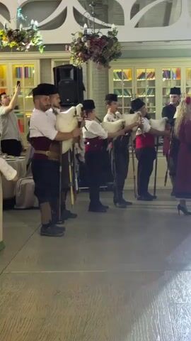 A group of men, dressed in traditional Bulgarian attire, are playing bagpipes in a room. They are standing in a line, facing to the right. The room is decorated with flowers and has a wooden floor. A woman in a colorful dress is standing to the right of the group, facing the men. There are bookshelves in the background.