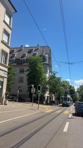 The video shows a tram moving down a city street on a sunny day. The tram is white and blue and has the number 7 displayed on its front. It passes by buildings and trees, and the street is lined with tram tracks and yellow pedestrian crossings. There are overhead wires for the tram. A traffic light is visible, and it is green. A car is also visible in the distance. The tram has a logo that says "VBZ Zürich Line".