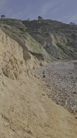 The video shows a person walking on a rocky beach towards the ocean. The person is wearing shorts and a light-colored shirt, and they appear to be carrying a bag or backpack. The beach is covered in large rocks and pebbles, and there is a steep, sandy cliff on the left side of the frame. In the distance, there are hills covered in green vegetation. The sky is clear and blue, suggesting a sunny day. The overall impression is of a remote and rugged coastline.