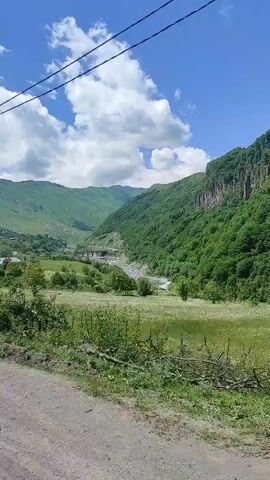 The video captures a scenic view of a lush green valley under a bright blue sky with fluffy white clouds. The camera appears to be mounted on a vehicle moving along a dirt road, evidenced by the slightly blurred foreground and the movement of the scenery.

The scene is dominated by rolling green mountains that rise steeply on either side of the valley. The slopes are densely covered with trees and vegetation. A winding river flows through the valley floor, and a bridge can be seen in the distance. The foreground consists of a rough dirt path with some scattered stones and dry grass. A wooden utility pole with wires runs vertically on the left side of the frame, indicating the presence of infrastructure in this natural setting.

The time of day appears to be late morning or early afternoon, given the bright sunlight casting clear shadows and illuminating the landscape. The weather is pleasant, with a clear sky suggesting a warm and possibly breezy day.

The location is Kvemo Mleta, Georgia, known for its mountainous terrain and natural beauty. The overall mood is tranquil and serene, highlighting the majestic landscape and the feeling of being in a remote, picturesque natural environment. No people are visible in the frame, and there is no visible text. The activity is the passive observation of the passing landscape from a moving vehicle.