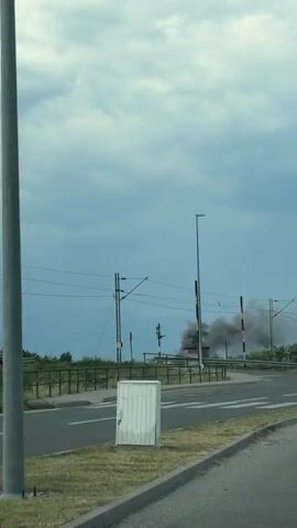 The video captures a scene in Goričani, Montenegro, with a prominent plume of dark smoke rising into the sky, indicating a possible fire or industrial event. The setting appears to be a roadside, with a clear view of a road, a pedestrian crossing, and power lines overhead, suggesting an urban or semi-urban environment.

In the foreground, the road with white lane markings and a pedestrian crossing is visible, indicating it's a public thoroughfare. A white directional arrow is painted on the road, pointing left. In the middle ground, there's a railway track with overhead electrical wires, and a small, simple building is situated near the tracks. A modern electrical cabinet is also present on the roadside. A black car is seen driving on the road.

The sky is overcast with dramatic, grey clouds, suggesting potentially unstable weather. The time of day appears to be daytime, given the ambient light. The smoke is the most significant element, dominating the background and suggesting an event of concern. The presence of trees and greenery suggests a developed area with some natural elements. There is no visible text in the video. The overall mood conveyed is one of concern or observation of an unusual event due to the heavy smoke.