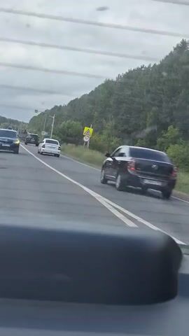 The video shows a view from inside a vehicle moving along a highway. Several cars are visible on the road, some in front and some in the opposite lane. The highway is lined with trees and a grassy embankment on the right side. The sky is overcast. Traffic appears to be moving normally.