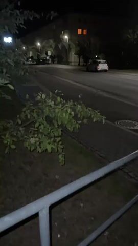 The video shows a nighttime street scene. The camera is positioned at ground level, looking across a paved area with grass and a metal railing in the foreground. Beyond the railing is a sidewalk and then a street. On the street, there are several cars. One white car is in motion, traveling away from the camera. Another dark-colored car is parked or moving slowly on the right side of the street. Streetlights illuminate the area, casting long shadows. In the background, there is a building with illuminated windows. The overall environment appears to be a residential or urban area at night.