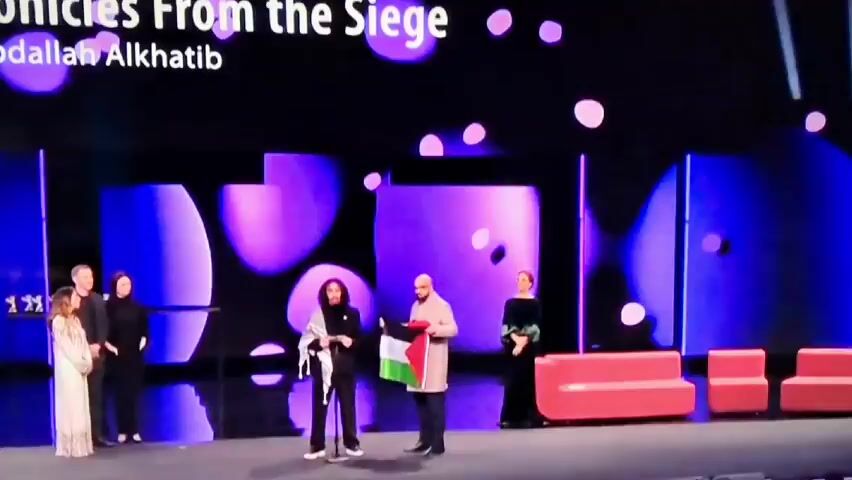 The video begins with a shot of a stage featuring several individuals.  One person in the center is holding a Palestinian flag. The stage is decorated with a backdrop of abstract shapes in shades of blue and purple.  There are also pieces of bright pink furniture on the stage.  The scene is captured from the perspective of an audience, suggesting a public event.  The video then cuts to a blurry dark shot before turning completely black.