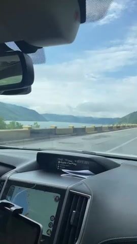 The video captures a scenic drive from the perspective of inside a car in Drobeta-Turnu Severin, Romania. The journey begins with a view of a wide road running parallel to a large body of water, likely a lake or river, bordered by distant, cloud-covered mountains. A concrete barrier with yellow pillars separates the road from the water. The sky is largely overcast with patches of blue visible.

Inside the car, parts of the dashboard and rearview mirror are visible. An infotainment screen displays "15:42" (time), "19.0" (possibly fuel or range), "Apple CarPlay," and temperature readings of "22.0°C" and "22°C." It also shows that the song "something about u." by "2Scratch" is playing. A lower screen, likely for navigation, is also visible.

As the video progresses, the view shifts to show the road winding alongside a green, mountainous slope. Other vehicles, including a dark SUV and several trucks, are seen driving ahead on the multi-lane road. The weather remains cloudy. The overall impression is a pleasant afternoon drive through picturesque natural landscapes.