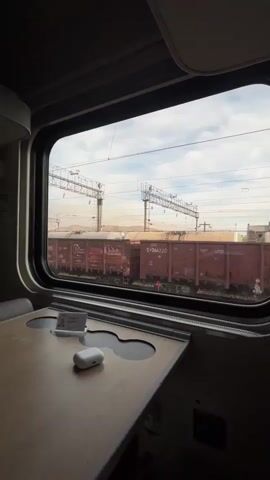 The video opens with a view from inside a train compartment, looking out a large rectangular window. A wooden tray table is visible in the foreground, with a small white item, likely earbuds, placed on it. To the left of the earbuds is a small, rectangular card. The window looks out onto a train yard. Several large, red freight cars are parked on the tracks. Overhead, power lines supported by metal structures stretch across the frame. The background shows a hazy, arid landscape with rolling hills. The sky is cloudy, with patches of blue visible. The camera pans slightly to the right, offering a closer view of the freight cars and the surrounding tracks.