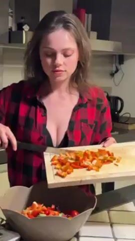 A woman is preparing food in a kitchen. She is wearing a red and black plaid flannel shirt over a black top. She is holding a wooden cutting board filled with diced orange and red vegetables. She uses a knife to push the vegetables off the cutting board into a brown bowl that already contains other chopped ingredients. A kettle is visible on the counter behind her, along with other kitchenware on a shelf.